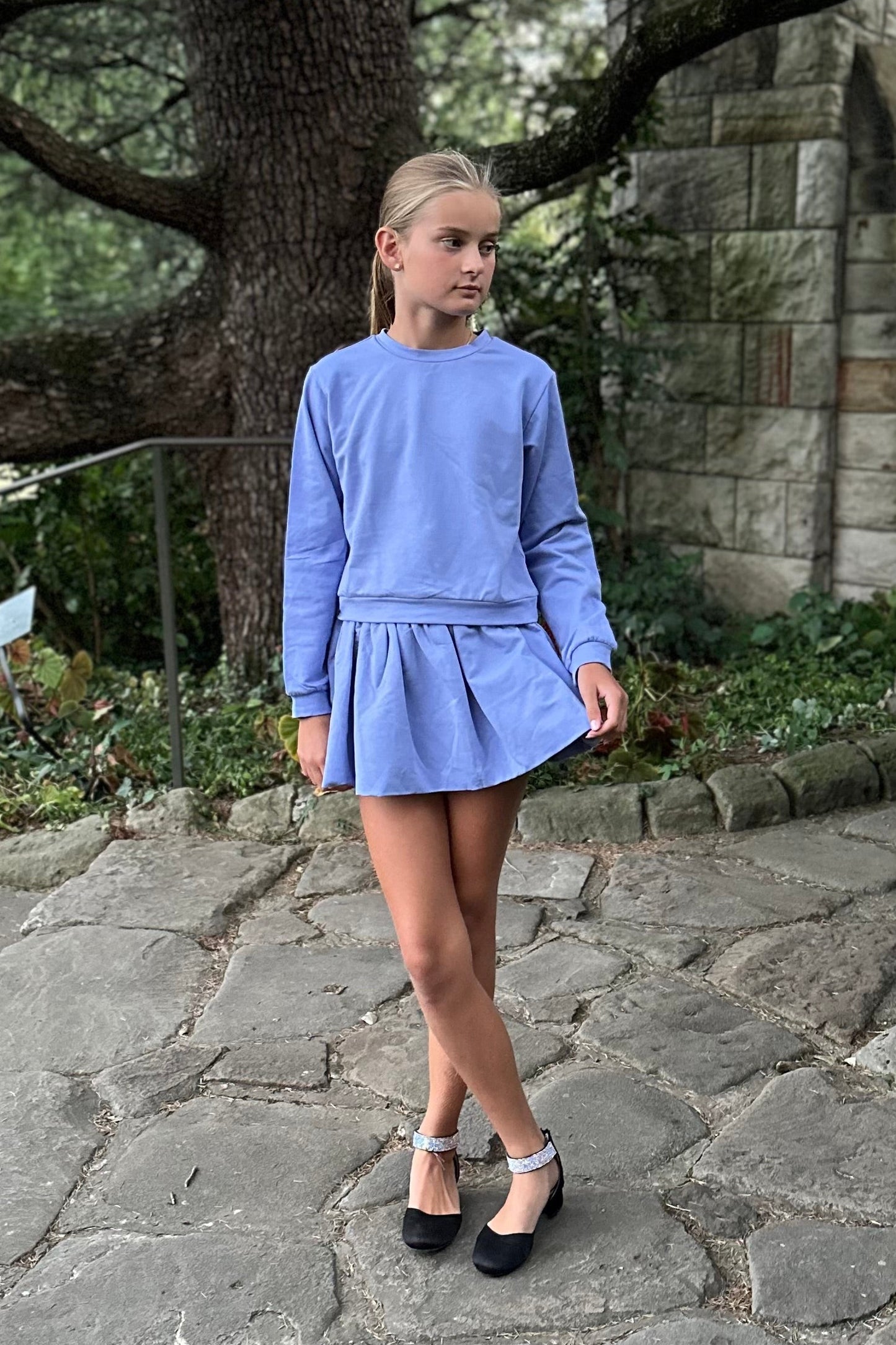 Person wearing a blue dress standing on a stone path with a stone wall and tree in the background